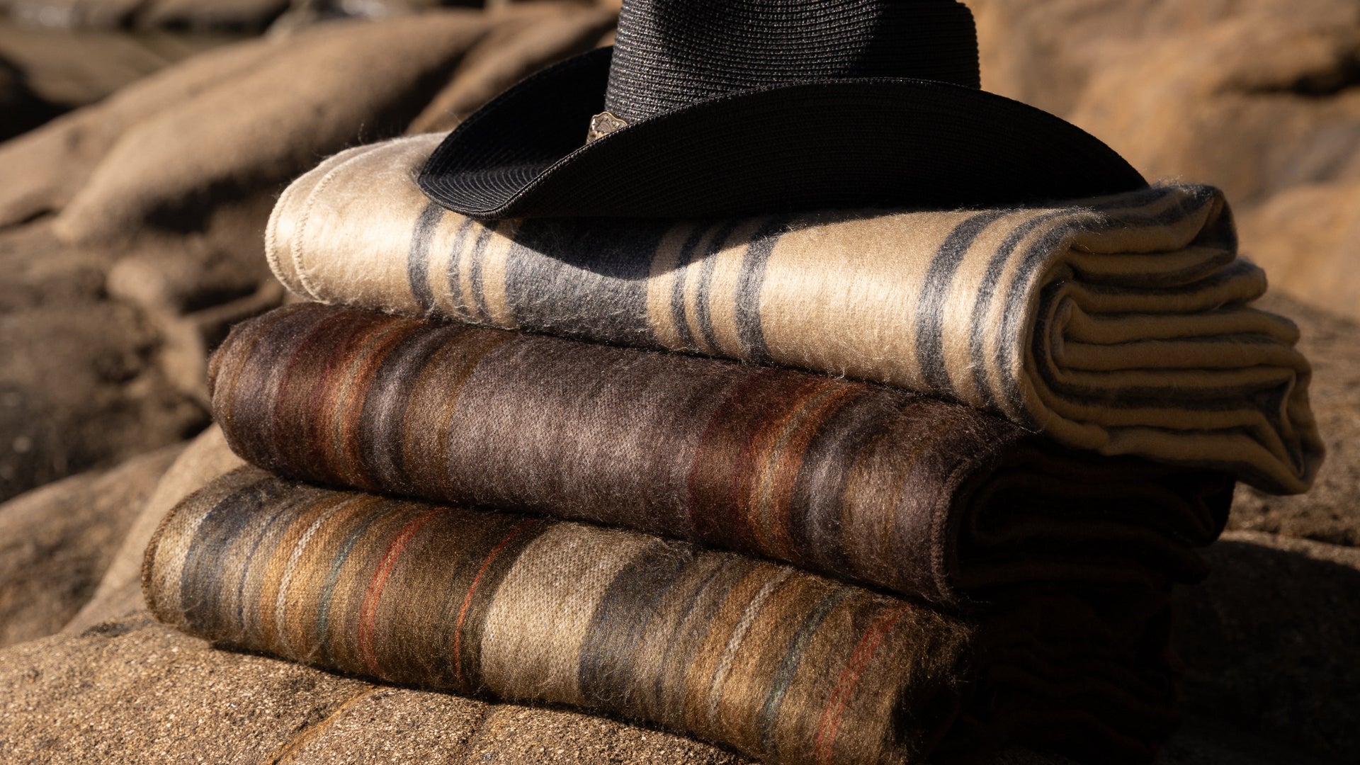 Stack of folded alpaca blankets and a black hat on a rocky surface with greenery in the background