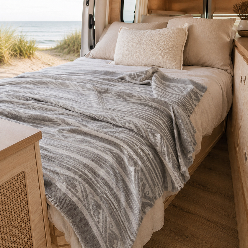 Bed in campervan with patterned llama wool blanket and pillows with beach view