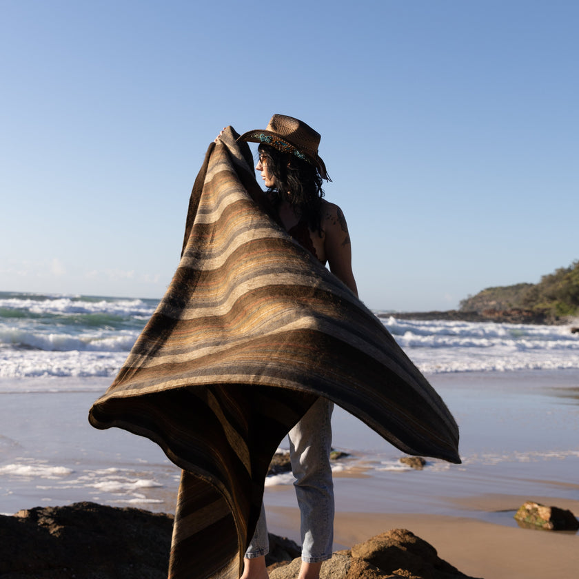 Person holding a large striped baby alpaca wool blanket on a rocky beach with ocean waves and clear sky.