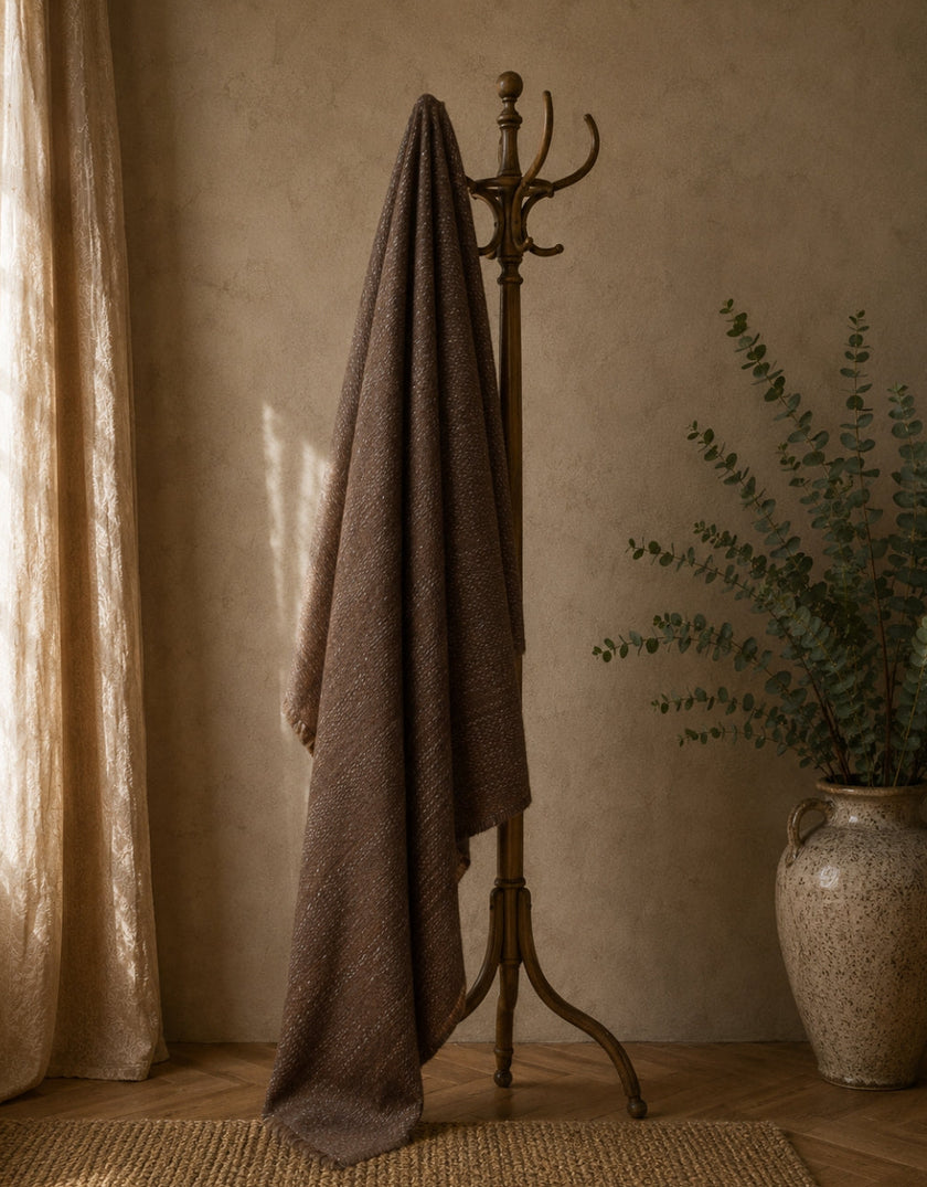 A dark brown textured alpaca wool blanket draped over a vintage bronze coat rack, set in a warm, rustic room with a textured beige wall, sheer curtains, and a potted eucalyptus plant.