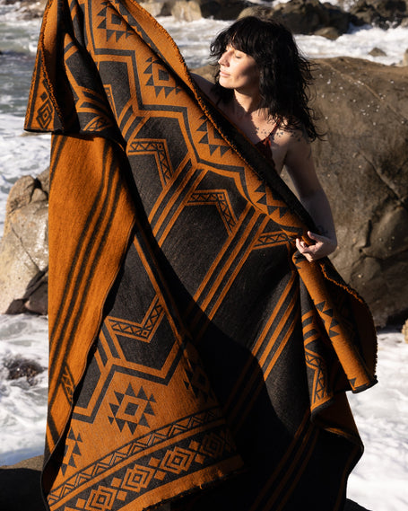 Person holding a patterned llama wool blanket by the ocean with rocky shores and greenery.