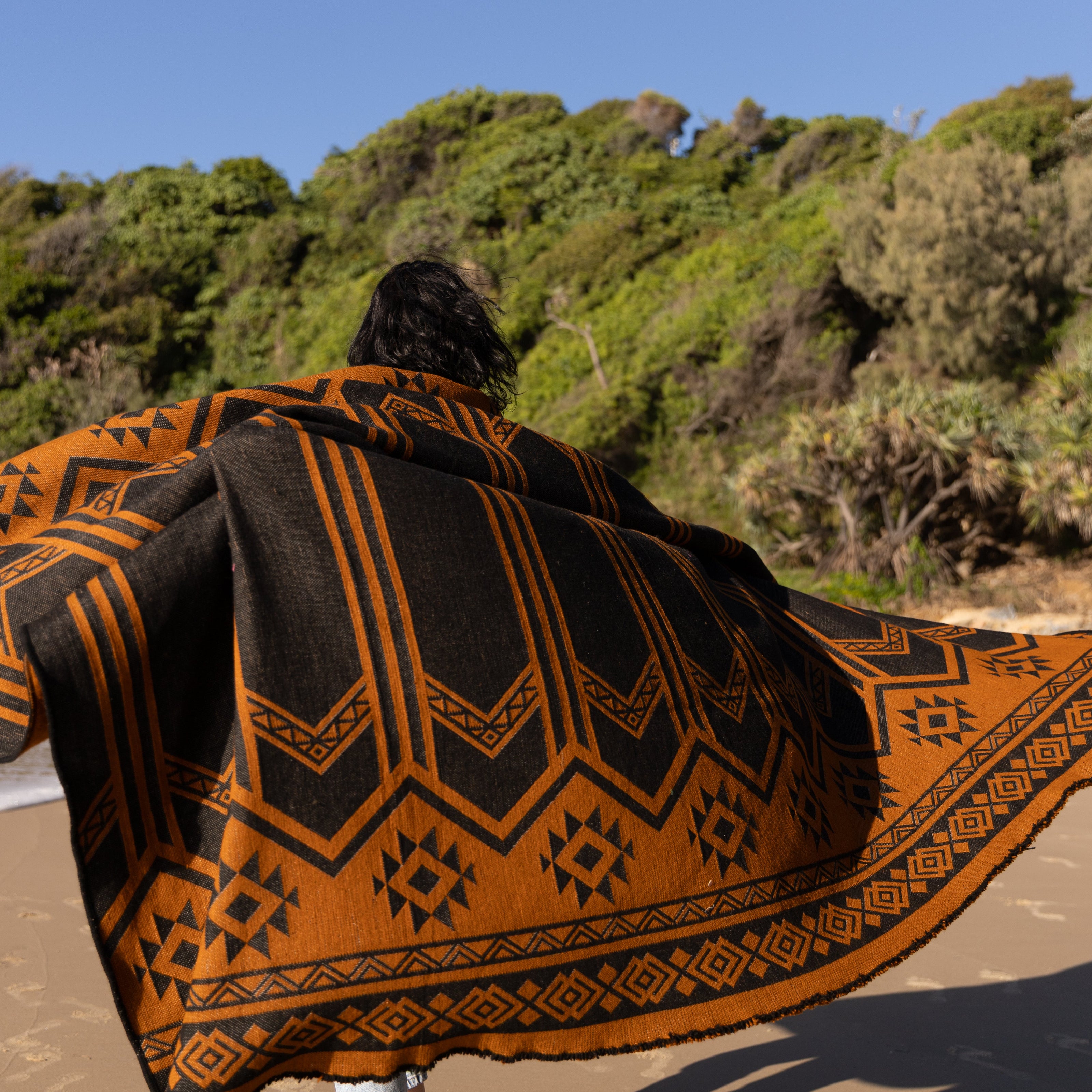 Person wearing a patterned llama wool blanket on an Australian beach with greenery in the background