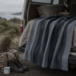 Van with bed setup outdoors, featuring a blue and grey alpaca blanket, pillows, and camping gear.
